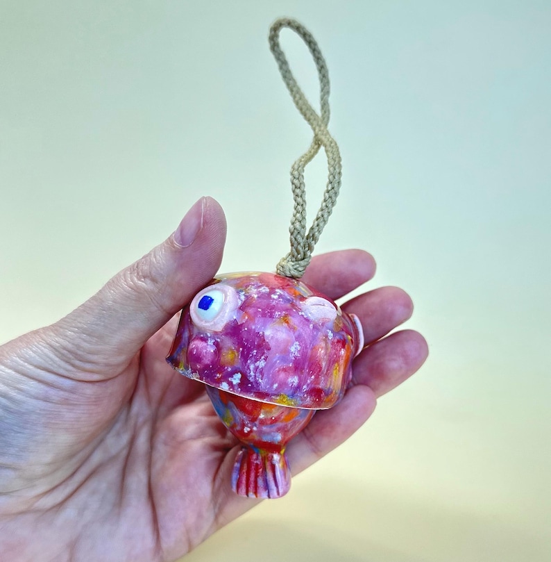 May include: A small, round, ceramic fish ornament featuring a vibrant, multi-colored glaze in shades of pink, purple, orange, and yellow. The fish has a single visible blue eye and a braided beige cord attached to the top for hanging purposes.