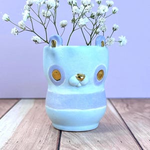 Puede incluir: Un jarrón de cerámica azul claro con forma de cara de oso, con detalles dorados en las orejas, la nariz y los ojos. El jarrón está lleno de flores blancas. Una franja morada clara rodea el centro del jarrón.