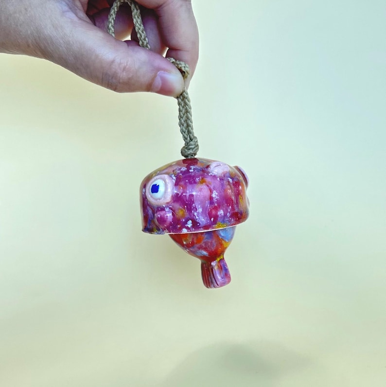 May include: A ceramic bell shaped like a pufferfish, featuring a glossy glaze in shades of pink, purple, and orange with white speckles. The fish has a prominent eye and a small tail, and it hangs from a thick, braided tan rope cord.