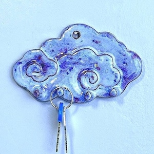 May include: A cloud-shaped key holder in shades of blue and purple. The ceramic piece has a glossy finish and a key ring with two keys attached. The design features swirling patterns and droplet-shaped details along the bottom edge.