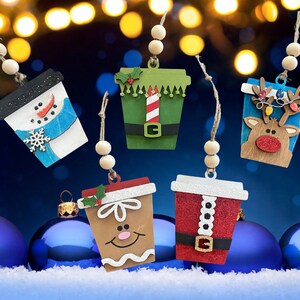 Puede incluir: Cinco adornos de madera con forma de vaso de caf&eacute; y motivos navide&ntilde;os: un mu&ntilde;eco de nieve, un elfo, un reno, un hombre de jengibre y un traje de Pap&aacute; Noel. Cada adorno incluye dos cuentas de madera y una cuerda de yute para colgar.