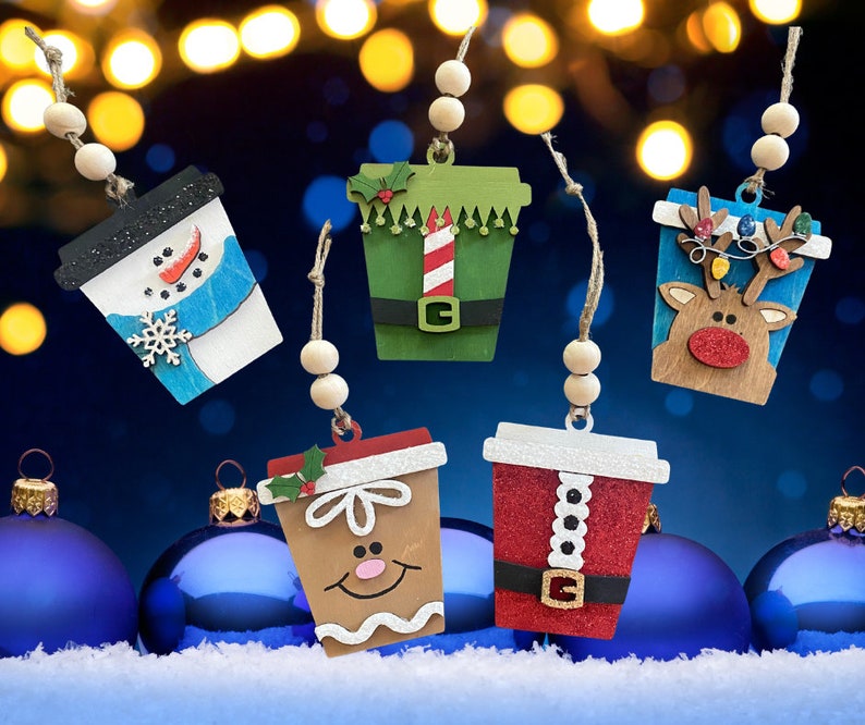 Puede incluir: Cinco adornos de madera con forma de vaso de caf&eacute; y motivos navide&ntilde;os: un mu&ntilde;eco de nieve, un elfo, un reno, un hombre de jengibre y un traje de Pap&aacute; Noel. Cada adorno incluye dos cuentas de madera y una cuerda de yute para colgar.