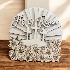 May include: Wooden Easter decoration with the words "He is Risen" in script. The design features three crosses, a rising sun, and a border of lilies. The piece is made of light-colored wood.