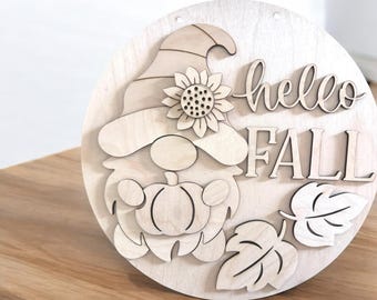 May include: A round wooden sign with a gnome design and the words "hello FALL". The gnome wears a hat with a sunflower and holds a pumpkin. Two leaves are also included in the design. The sign is made of light-colored wood.