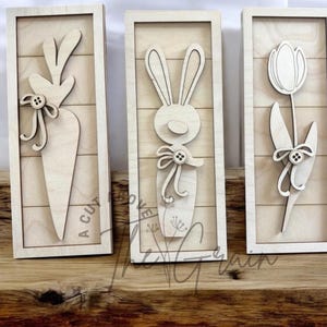 May include: Three wooden signs with a farmhouse style design. The signs feature a carrot, a bunny rabbit, and a tulip. Each sign has a white background with a light brown wood grain texture.