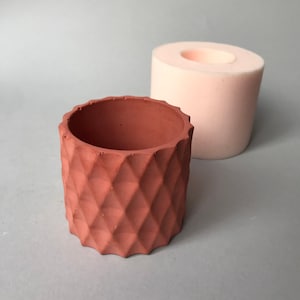 Può includere: Un vaso color terracotta, con motivo a diamante e superficie strutturata. Accanto, uno stampo cilindrico rosa chiaro. Il vaso ha un bordo ruvido e scanalato. Ideale per piccole piante o succulente.