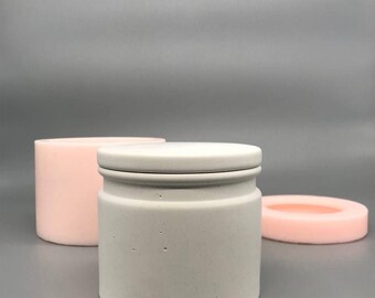 May include: A white concrete jar with a lid, sitting on a gray surface. The jar has a rough texture and is slightly wider at the top than the bottom. The lid is also made of concrete and has a smooth surface.