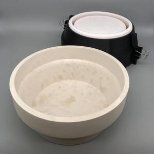 Può includere: Due stampi rotondi in ceramica bianca per realizzare vasi in cemento. Lo stampo più grande è appoggiato su un tavolo. Lo stampo più piccolo è all'interno dello stampo più grande. Lo stampo più piccolo ha un anello di plastica nera attorno alla parte superiore.