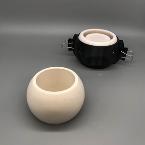 May include: A spherical off-white ceramic vessel with an open top sits next to a black 3D-printed mould assembly. The mould consists of a white inner core held within a black outer shell, secured by two silver metal binder clips on the sides.