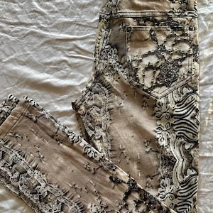 Może przedstawiać: Dżinsy w brązowo-białe wzory z motywem kwiatowym. Dżinsy mają jasnobrązowy kolor tła z białym wzorem kwiatowym.