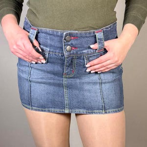 May include: A denim mini skirt with a classic five-pocket design. The skirt has a button and zip closure, with red stitching accents. The garment is worn with a long-sleeved olive green top.