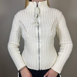 May include: Cream-colored ribbed knit zip-up sweater with a high collar. The sweater features a vertical ribbed design, a full-length zipper, and decorative cutouts near the shoulders. The sleeves are long, and the sweater has a fitted silhouette.