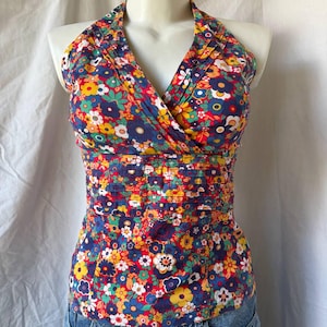 May include: A sleeveless halter top featuring a vibrant floral pattern with red, yellow, blue, and green flowers on a dark blue background. The top has a V-neckline with pleated detailing at the bodice and a wide, horizontally pleated waistband.