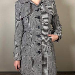 May include: A black and white geometric patterned coat with a notched collar, long sleeves, and a button-down front. The coat features a unique design with black buttons and pockets.