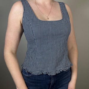 May include: A navy and white checked sleeveless top with a square neckline and a frilled hem. The top is worn with dark blue jeans. A gold necklace is also visible.