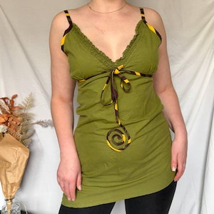 Op de afbeelding: Olijfgroene top met spaghettibandjes, een V-hals en een decoratief spiraalontwerp. De bandjes en halslijn zijn afgewerkt met een bruin en geel gestreept patroon. De top wordt gecombineerd met een zwarte legging.