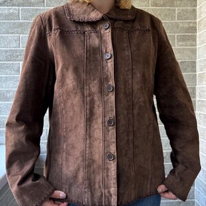 May include: A brown suede jacket with a button-up front and a collared neckline. The jacket features vertical stitched detailing and decorative stitching along the collar. The sleeves are long, and the jacket is worn over blue jeans.