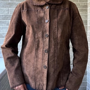 May include: A brown suede jacket with a button-up front, a rounded collar, and decorative stitching. The jacket has long sleeves and vertical seams down the front. The jacket is worn over blue jeans.