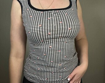 May include: A sleeveless top with a black and white gingham pattern and a square neckline. The top has a black trim and small pink floral accents scattered across the fabric. The garment is made of a soft, form-fitting material.