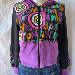 Puede incluir: Una sudadera con cremallera que presenta una sección superior negra con texto colorido estilo grafiti y parches circulares rosas, blancos y amarillos. La sección inferior y los puños son morados, mientras que las mangas son negras con detalles morados y naranjas.