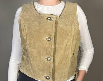 Puede incluir: Chaleco de ante color canela con cierre de botones y bordados decorativos. El chaleco tiene un escote redondeado y un corte entallado. Los botones son plateados. Se lleva sobre una camisa blanca de manga larga y pantalones negros.