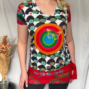May include: A V-neck t-shirt with a colorful, geometric pattern and a large, embroidered butterfly. The shirt features a red waist tie and is paired with dark gray leggings. The design incorporates circles and a rainbow color scheme.