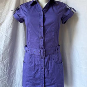 May include: A solid purple button-down shirt dress featuring short sleeves with buckled tabs, a classic collar, and a matching fabric belt with a rectangular buckle. The garment includes two front patch pockets and a full-length button placket.