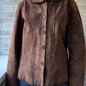 May include: A brown suede jacket with a rounded collar and button-up front. The jacket features decorative stitching along the collar, seams, and hem. The jacket has long sleeves and is made of a soft, textured material.