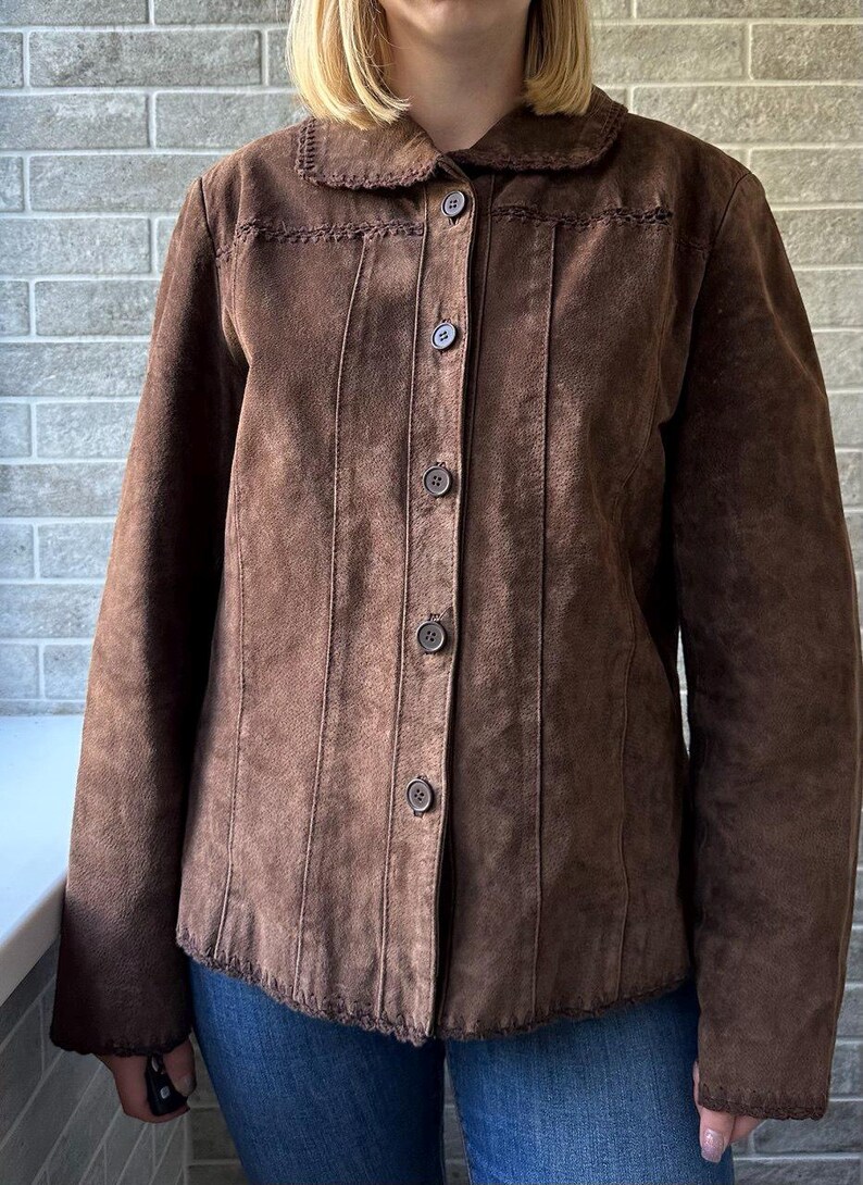 May include: A brown suede jacket with a rounded collar and button-up front. The jacket features decorative stitching along the collar, seams, and hem. The jacket has long sleeves and is made of a soft, textured material.