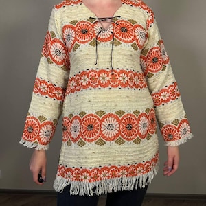 Puede incluir: Túnica color crema con mangas largas y cuello en V. La túnica presenta un estampado floral en naranja y verde oliva, con dobladillo y extremos de manga con flecos. El escote tiene un detalle de cordones negros.