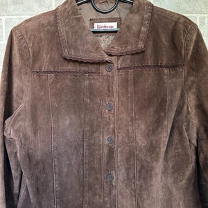 May include: A brown suede jacket with a collared neckline and button-down front. The jacket features decorative stitching along the collar and seams, and the label reads "Kaleidoscope". The jacket is hanging on a black hanger.