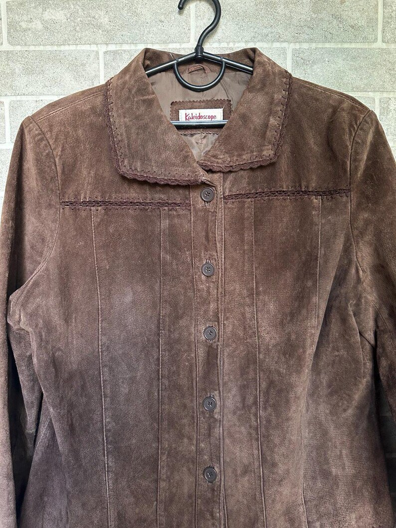 May include: A brown suede jacket with a collared neckline and button-down front. The jacket features decorative stitching along the collar and seams, and the label reads "Kaleidoscope". The jacket is hanging on a black hanger.