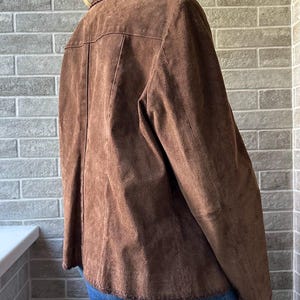 May include: A brown suede jacket with a collared neckline and button closure. The jacket has a tailored fit and features decorative stitching along the collar and hem. The jacket is worn over blue jeans.