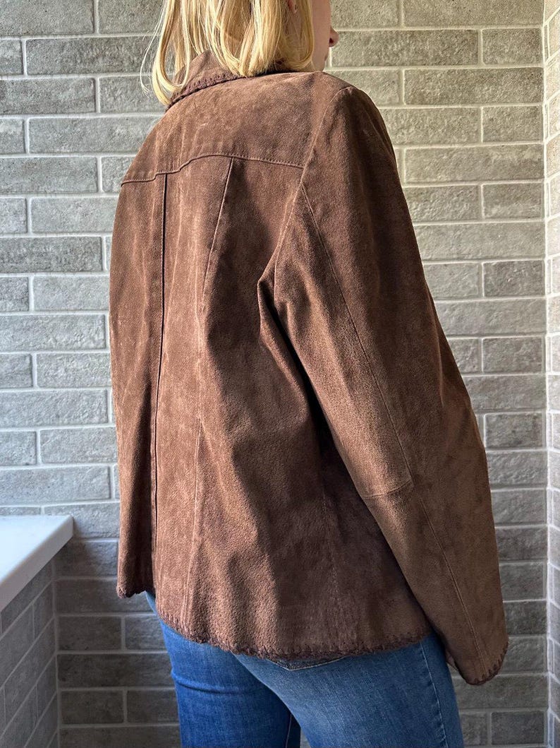 May include: A brown suede jacket with a collared neckline and button closure. The jacket has a tailored fit and features decorative stitching along the collar and hem. The jacket is worn over blue jeans.