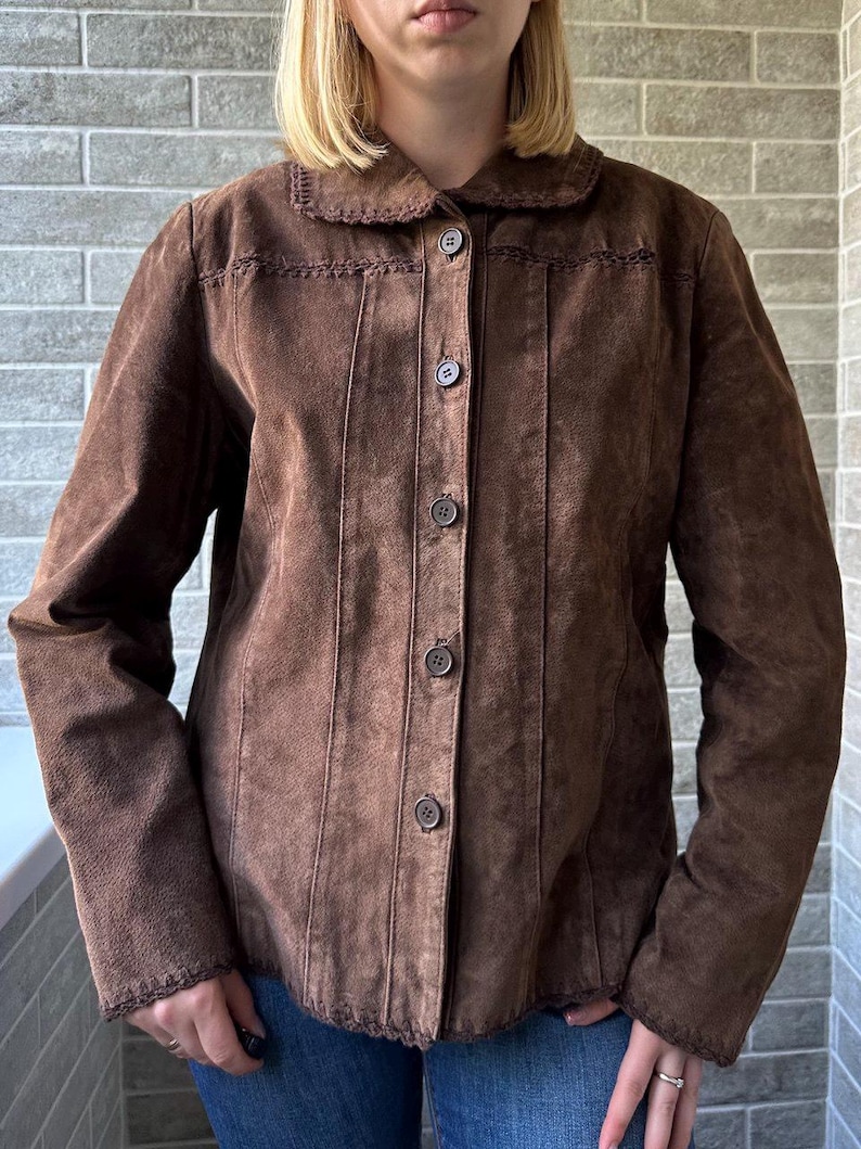 May include: A brown suede jacket with a button-up front, a rounded collar, and decorative stitching. The jacket has long sleeves and vertical seams down the front. The jacket is worn over blue jeans.