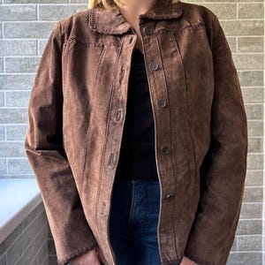 May include: A brown suede jacket with a button-up front and a collared neckline. The jacket features decorative stitching along the collar and seams. The model is wearing a black top and blue jeans.