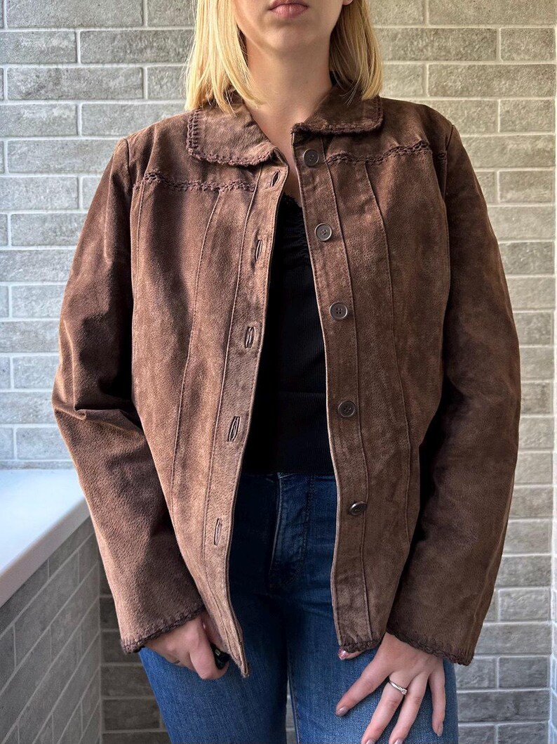 May include: A brown suede jacket with a button-up front and a collared neckline. The jacket features decorative stitching along the collar and seams. The model is wearing a black top and blue jeans.