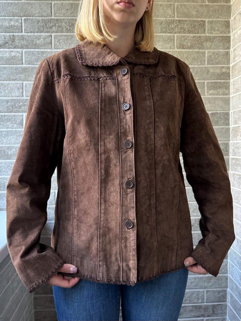 May include: A brown suede jacket with a button-up front and a collared neckline. The jacket features vertical stitched detailing and decorative stitching along the collar. The sleeves are long, and the jacket is worn over blue jeans.