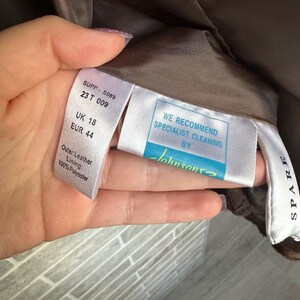 May include: Close-up of garment tags with text. The tags include size information: UK 18, EUR 44. One tag reads "Outer: Leather, Lining: 100% Polyester." Another tag recommends specialist cleaning by "Johnsons."