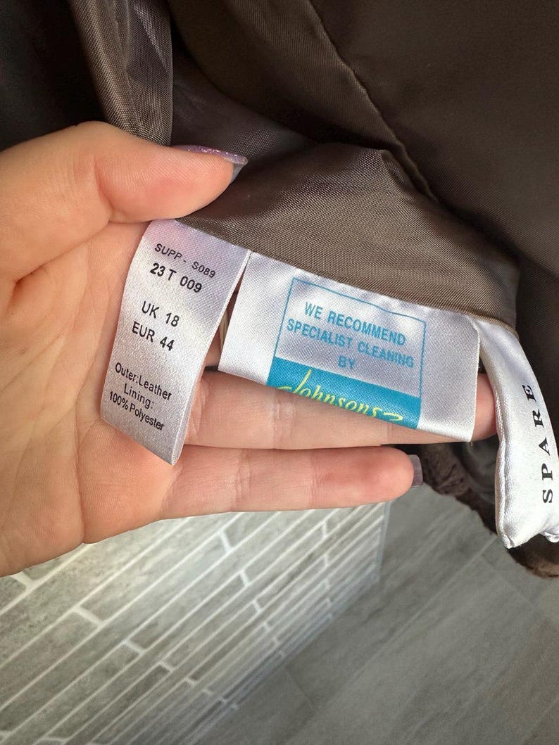 May include: Close-up of garment tags with text. The tags include size information: UK 18, EUR 44. One tag reads "Outer: Leather, Lining: 100% Polyester." Another tag recommends specialist cleaning by "Johnsons."