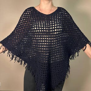 May include: A navy blue crochet poncho with a loose, open weave design. The poncho has a wide neckline and fringed edges along the bottom. The garment is made of a soft, lightweight material.