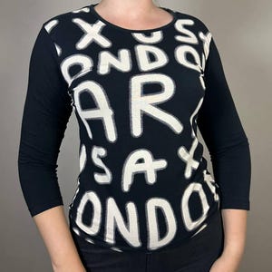 May include: Black long-sleeve shirt with a crew neck. The shirt features a bold white text design that reads "LONDON" and "SAX". The sleeves are three-quarter length. The shirt is made of a soft, comfortable material.