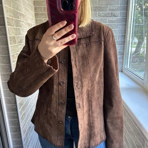 May include: A brown suede jacket with a button-up front and a decorative stitched collar. The jacket has long sleeves and is worn over a black top and blue jeans. The jacket is a medium brown colour.
