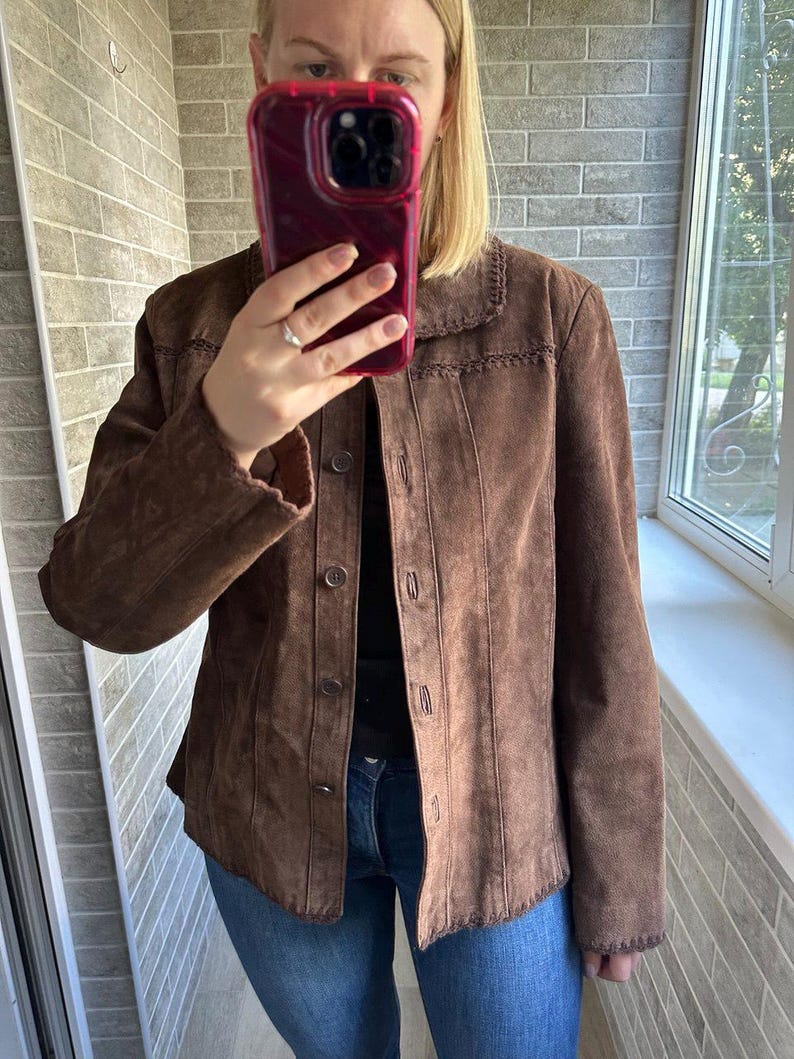 May include: A brown suede jacket with a button-up front and a decorative stitched collar. The jacket has long sleeves and is worn over a black top and blue jeans. The jacket is a medium brown colour.