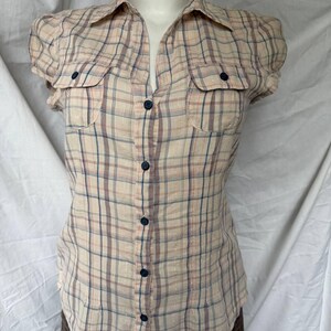 Peut inclure: Une chemise à manches courtes boutonnée avec un motif à carreaux beige, bleu et marron. La chemise présente un col classique, deux poches de poitrine avec des boutons bleu foncé et une fermeture boutonnée sur toute la longueur devant.