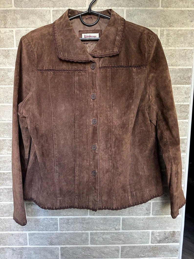 May include: A brown suede jacket with a button-up front and a collar. The jacket features decorative stitching along the seams and around the collar and hem. The label reads "Kaleidoscope".