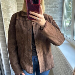 May include: A brown suede jacket with a button-up front and a collared neckline. The jacket features decorative stitching along the collar and seams. The model is wearing blue jeans.