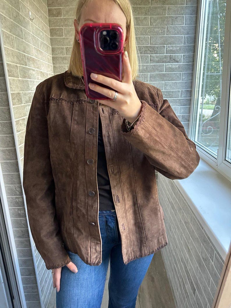 May include: A brown suede jacket with a button-up front and a collared neckline. The jacket features decorative stitching along the collar and seams. The model is wearing blue jeans.
