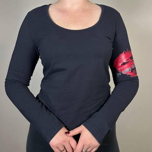 May include: A dark grey long-sleeved top with a scoop neckline. A red lip print is on the left sleeve. The shirt is made of a soft material and has thumb holes at the cuffs. A casual top for everyday wear.