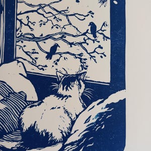 May include: A blue and white linocut print depicting a cat sitting on a surface, looking out a window at two birds perched on tree branches. The print features the text "Vue sur le jardin" and initials at the bottom, rendered in a high-contrast style.